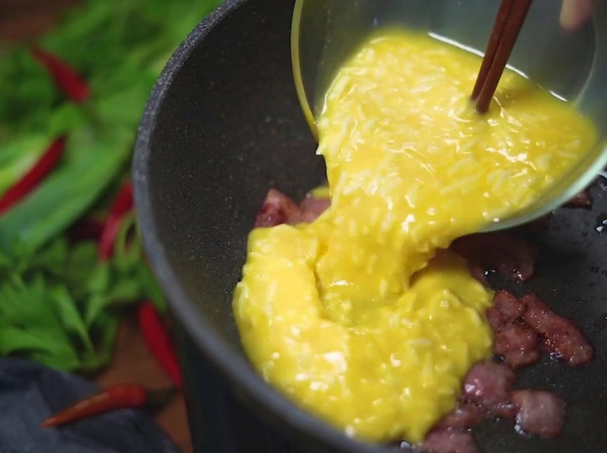 Un composto giallo di uova e formaggio versato da una ciotola di vetro in una padella calda contenente pezzetti di bacon croccanti e dorati.