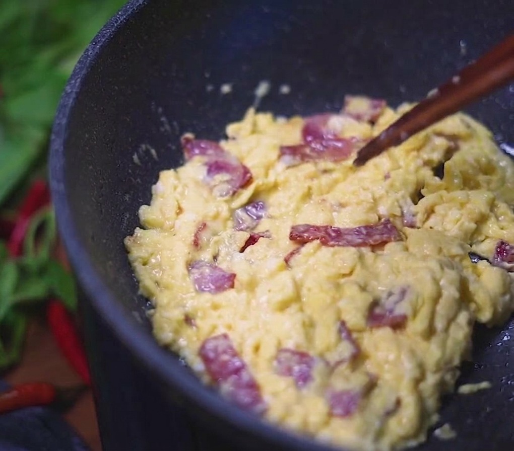 Uova strapazzate cremose mescolate con pezzetti di bacon saporiti, girate in una padella scura con delle bacchette.
