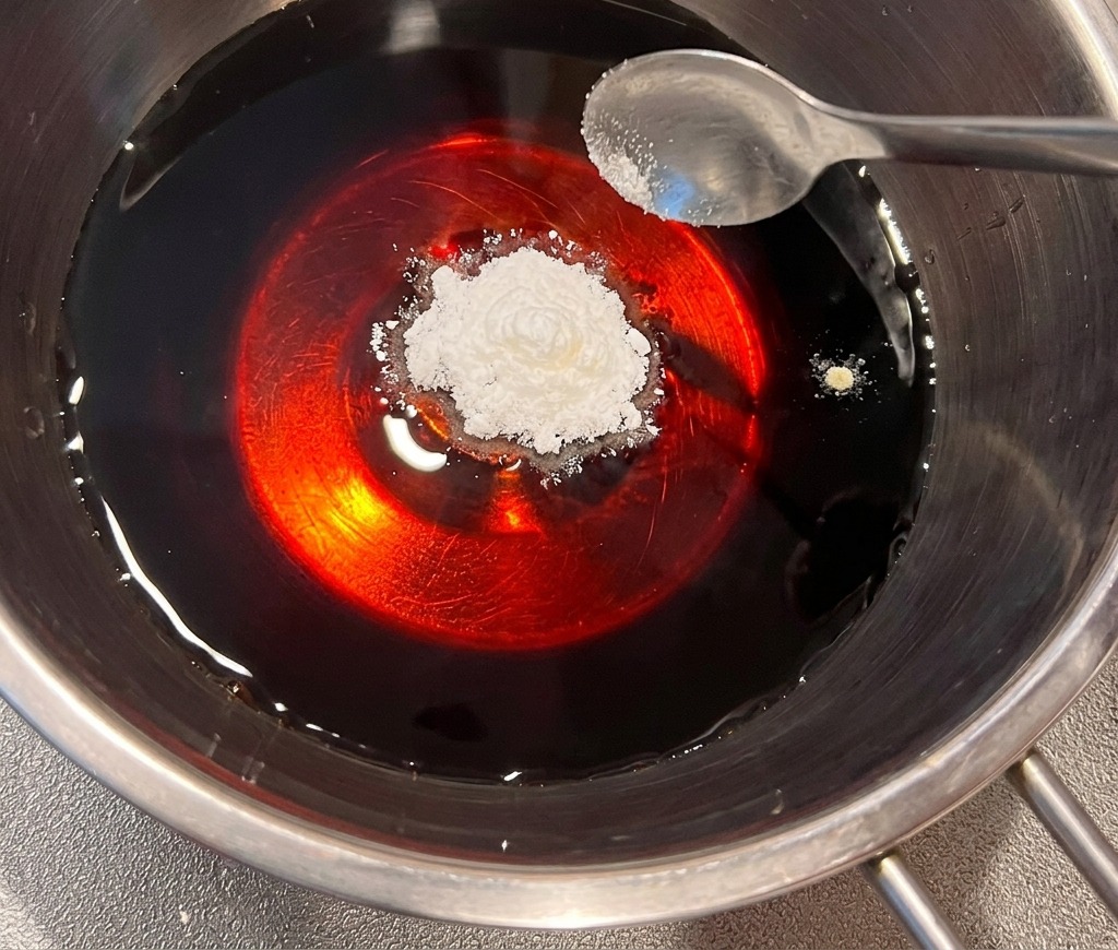 Un cucchiaio di polvere di dashi bianca viene aggiunto a un liquido scuro a base di soia in una pentola di acciaio inossidabile.