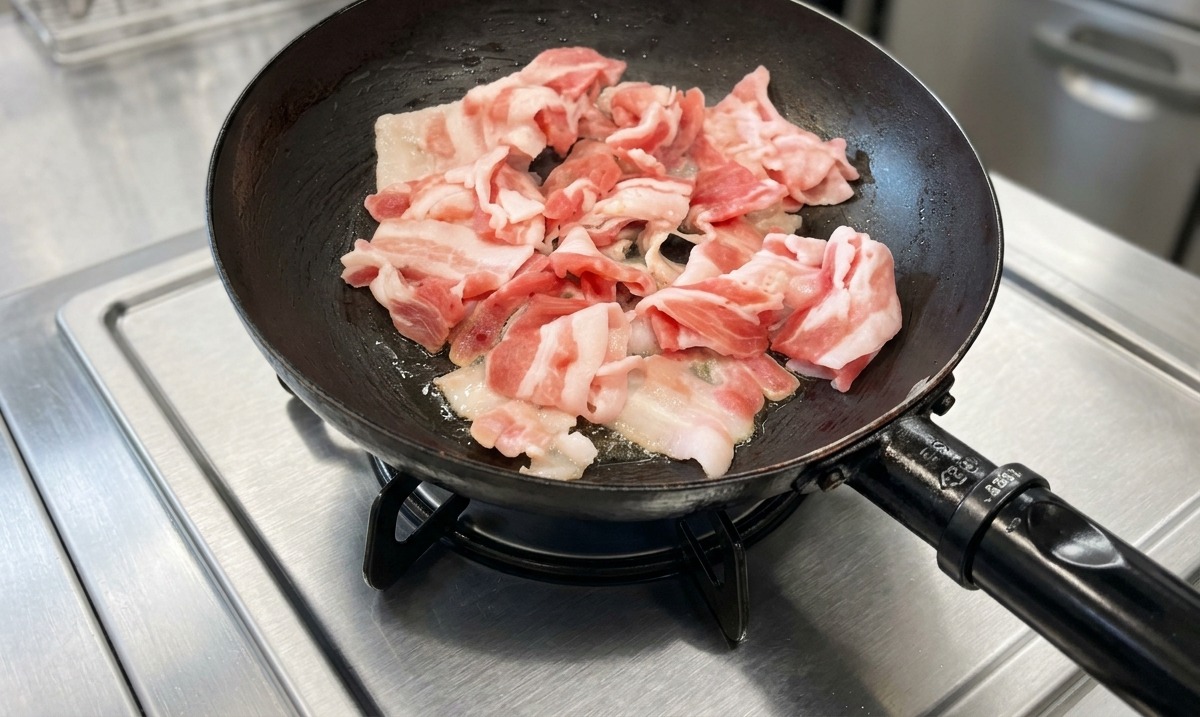 Fette di pancetta di maiale in cottura in una padella nera su un piano cottura in acciaio inossidabile.