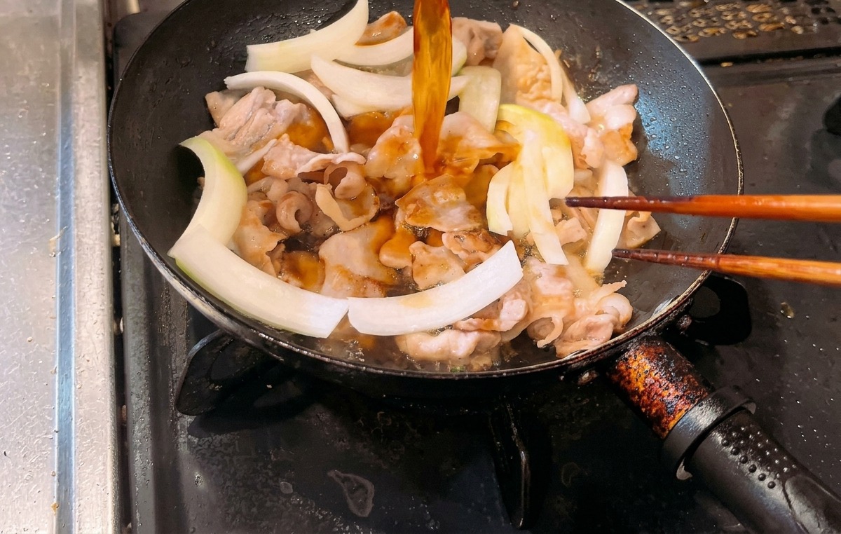 Versamento di una salsa color ambra in una padella contenente pancetta di maiale cotta e fette di cipolla bianca cruda.