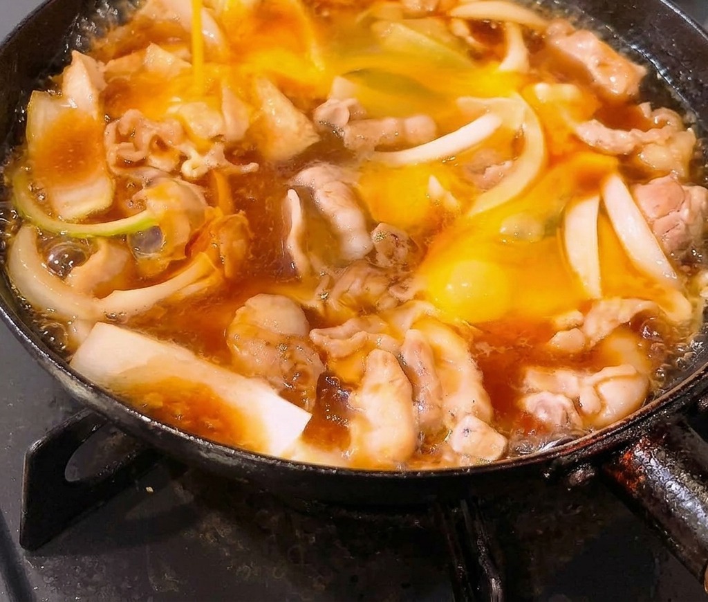 Uova sbattute versate in una padella piena di pancetta di maiale e cipolle che sobbollono in una salsa a base di dashi.