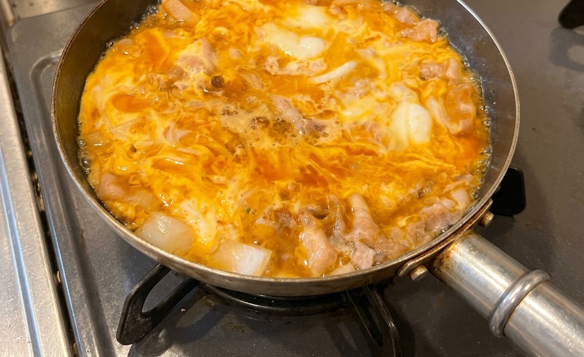 Una padella di Tanindon che sobbolle sul fuoco, con le uova gialle che diventano opache e rapprese attorno al maiale e alle cipolle.