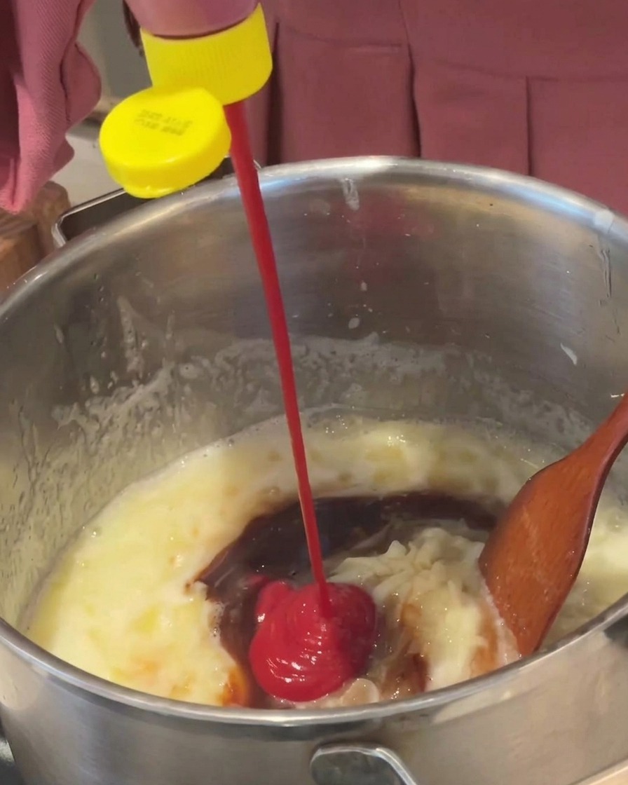 Ketchup rosso brillante spremuto da una bottiglia con tappo giallo in una salsa marrone che sobbolle in una pentola di metallo.