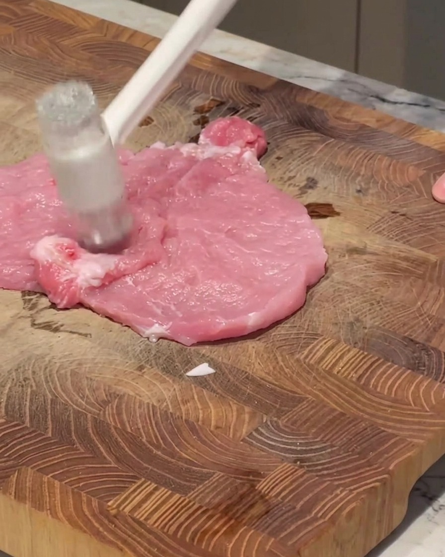 Un batticarne bianco che batte un pezzo di lonza di maiale cruda e rosa su un tagliere di legno.