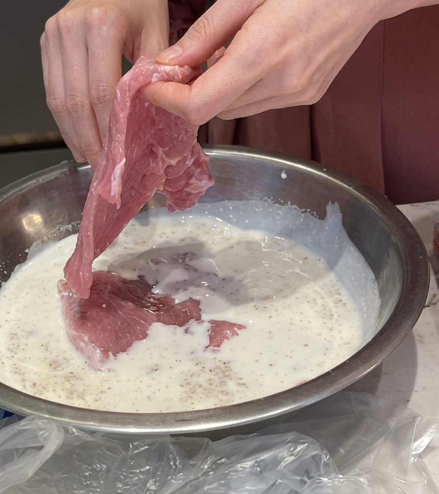 Mani che immergono una sottile fetta di maiale rosa in una ciotola di metallo piena di marinatura bianca a base di latte e aglio.
