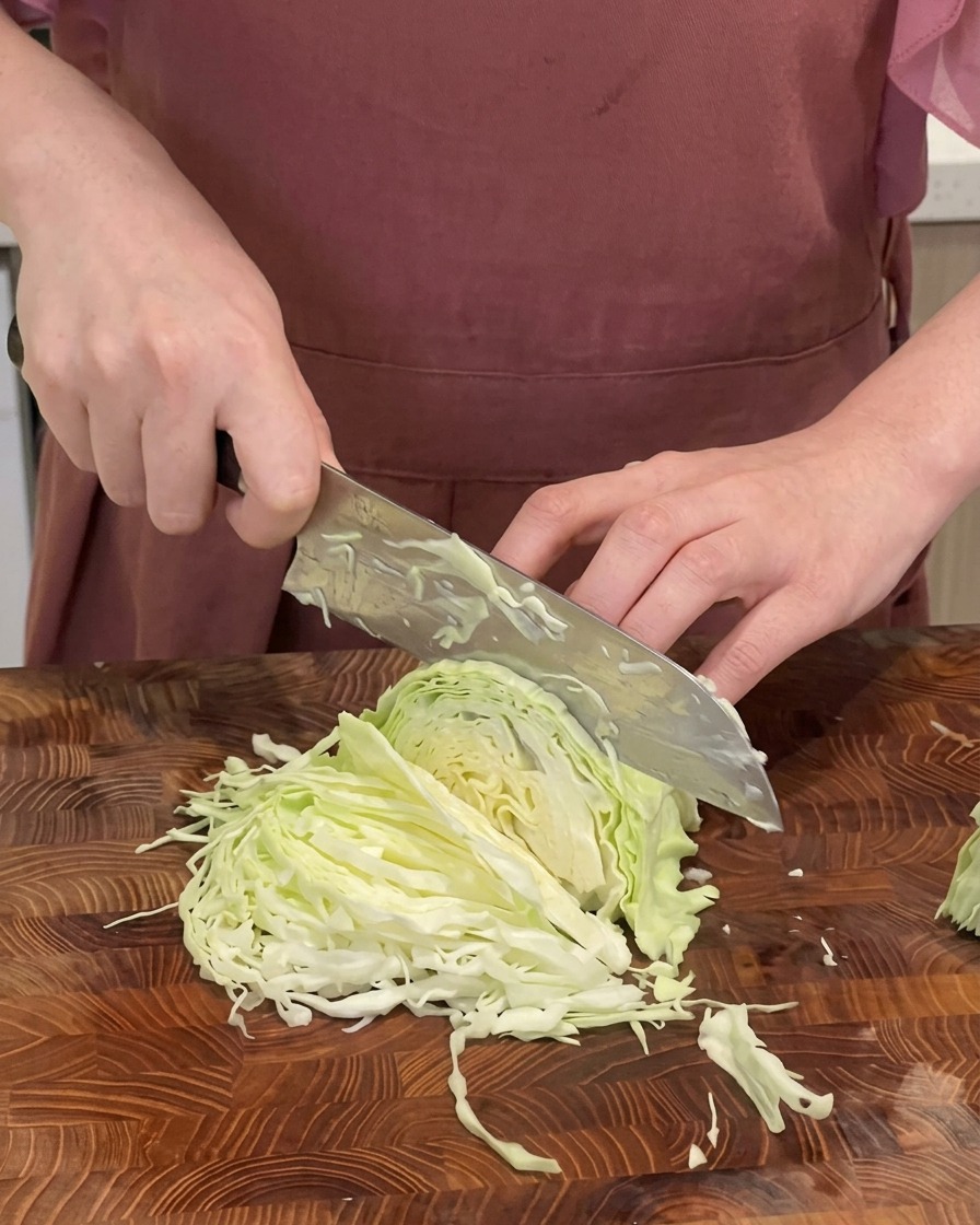 Primo piano di uno chef che sminuzza un cavolo cappuccio verde in strisce sottili su un tagliere di legno con un grande coltello.