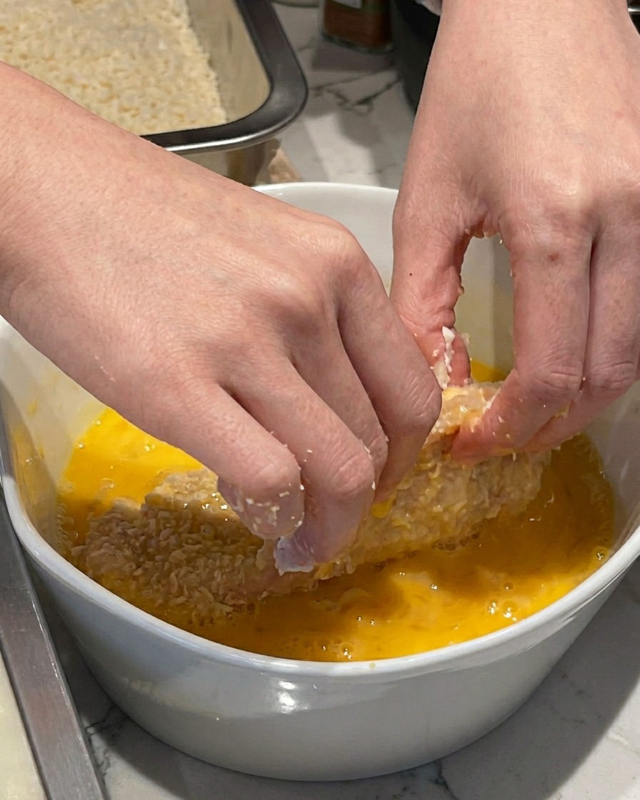 Una mano che immerge una cotoletta infarinata in una ciotola di ceramica bianca con uova sbattute.