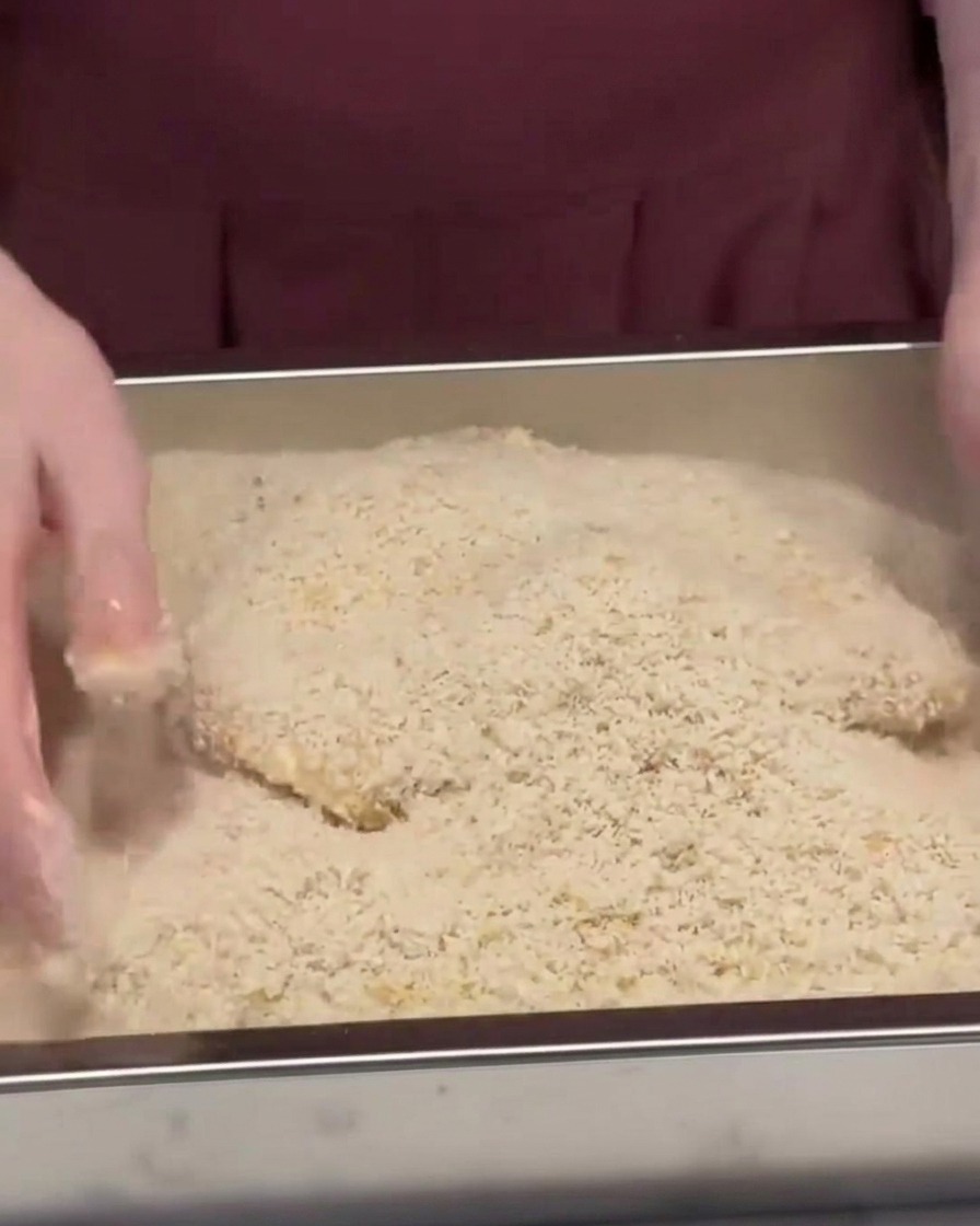 Una cotoletta di maiale coperta e pressata in un vassoio di metallo pieno di pangrattato panko.