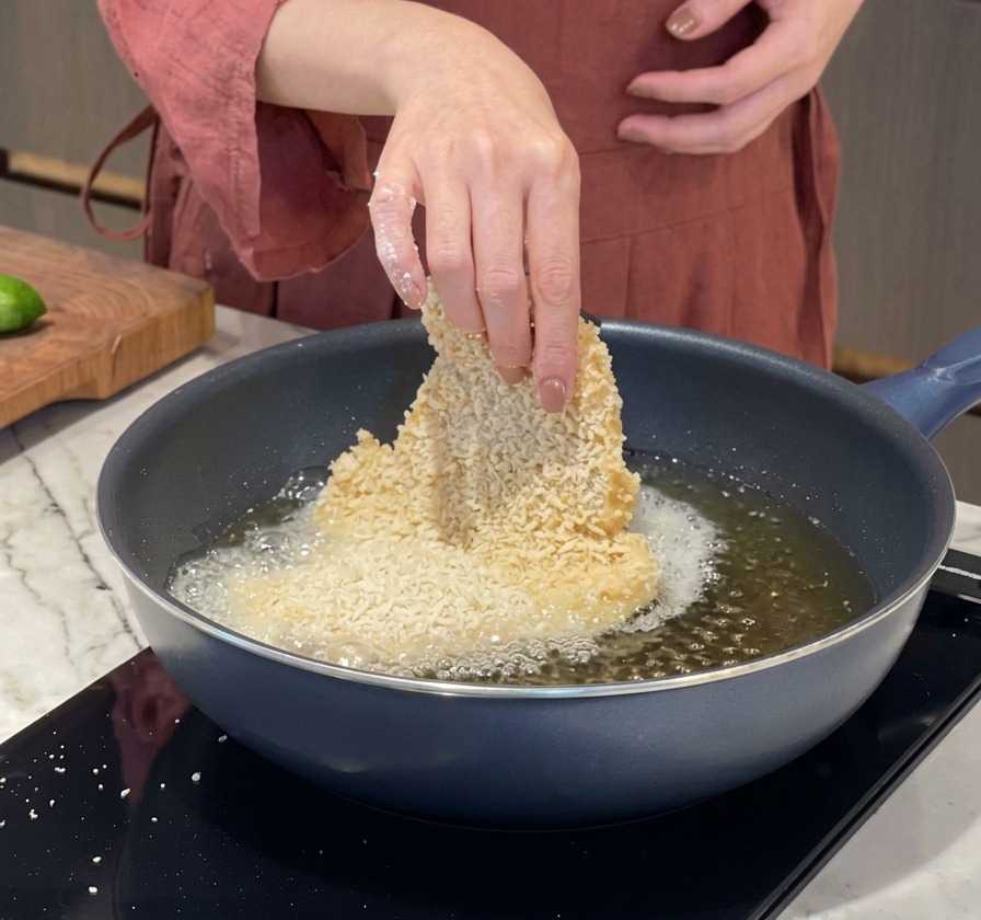 La mano di una persona che immerge delicatamente una grande cotoletta impanata in una padella calda con olio bollente.