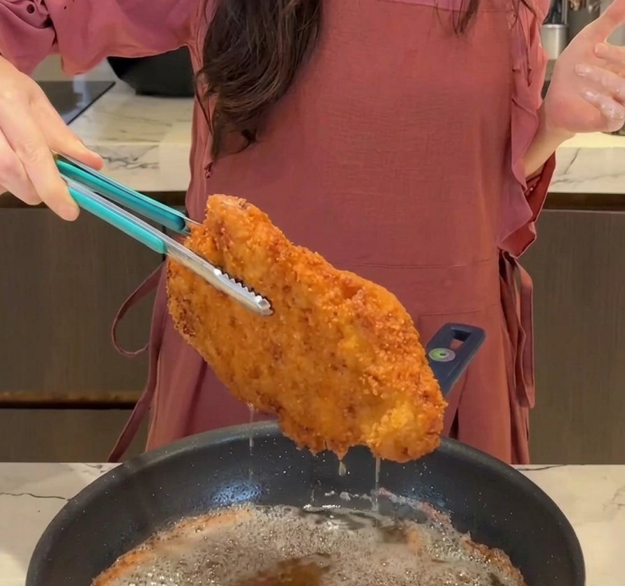 Una grande cotoletta di maiale fritta croccante e dorata sollevata da una padella con pinze blu, con lolio che gocciola sul fondo.