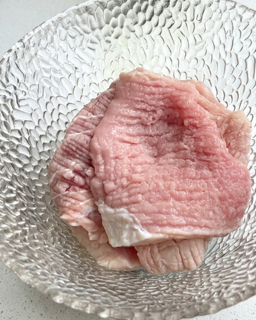Un primo piano di una ciotola di vetro contenente fette di maiale crudo condite con cipollotti tritati e aromi liquidi.
