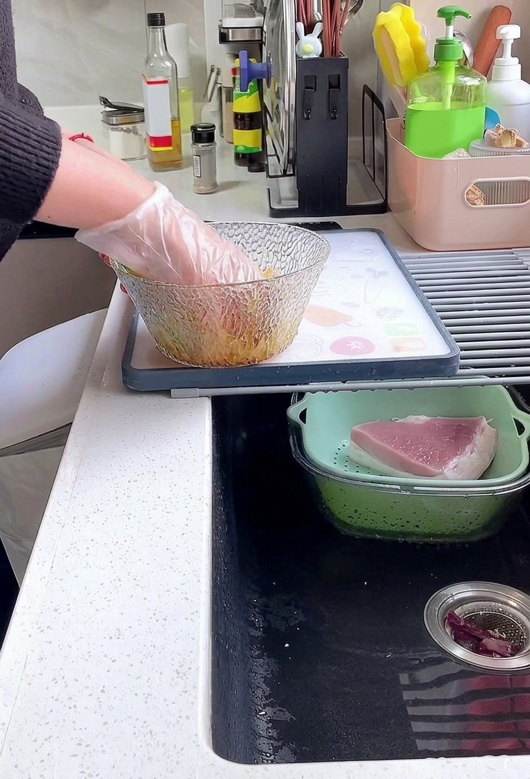 Una persona che indossa un guanto di plastica mescola spesse fette di lonza di maiale con salsa di soia, pepe e amido in una ciotola di vetro.