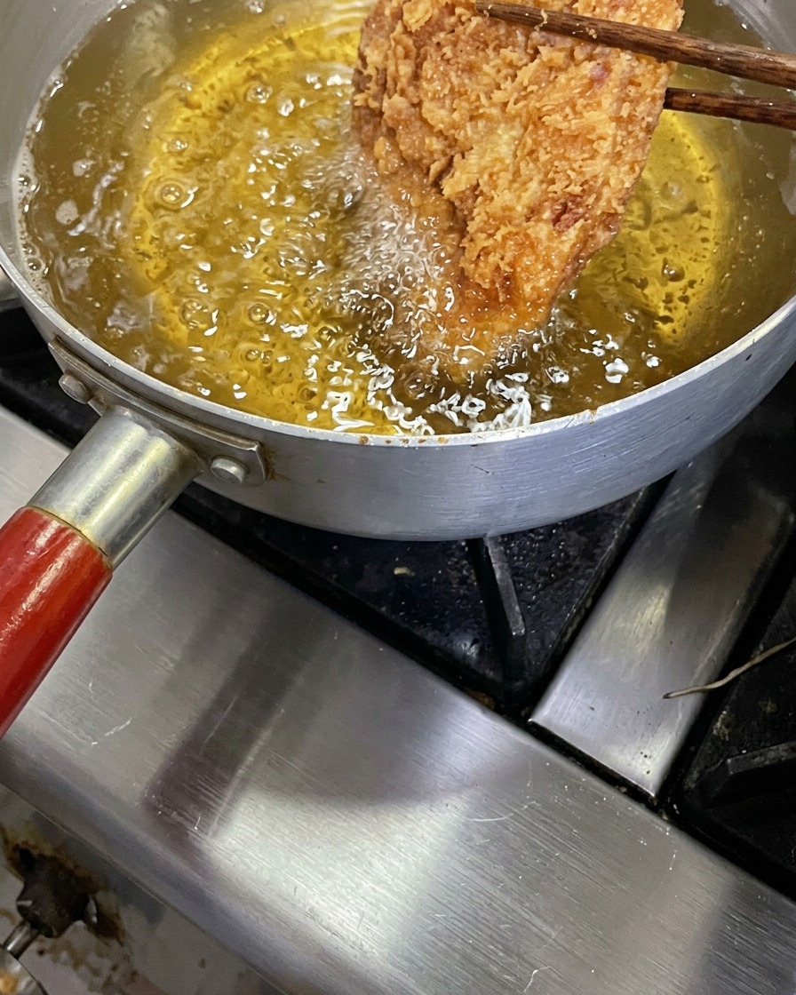 Una cotoletta tonkatsu dorata e croccante sollevata con bacchette di legno da una pentola di olio bollente.