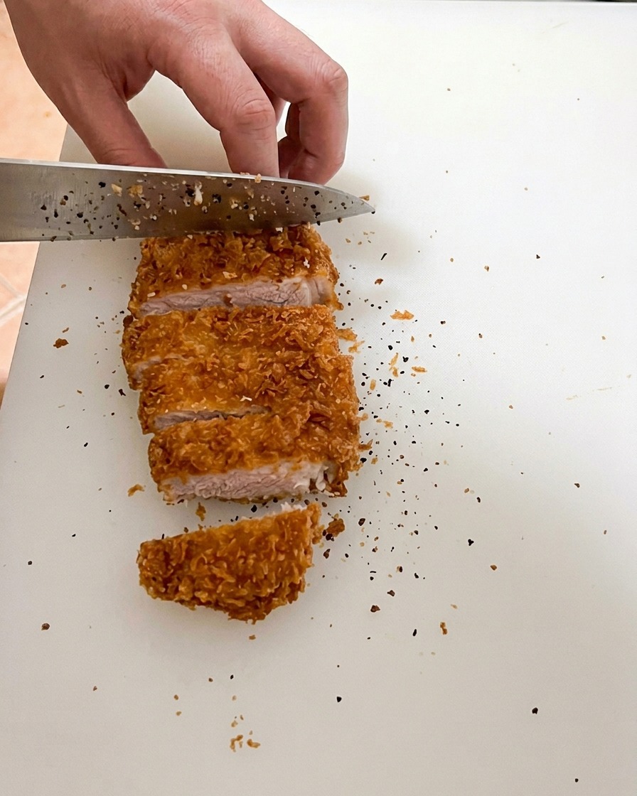 Un coltello affilato che taglia una cotoletta di maiale perfettamente fritta su un tagliere bianco, mostrando la carne succosa con un interno leggermente rosato.
