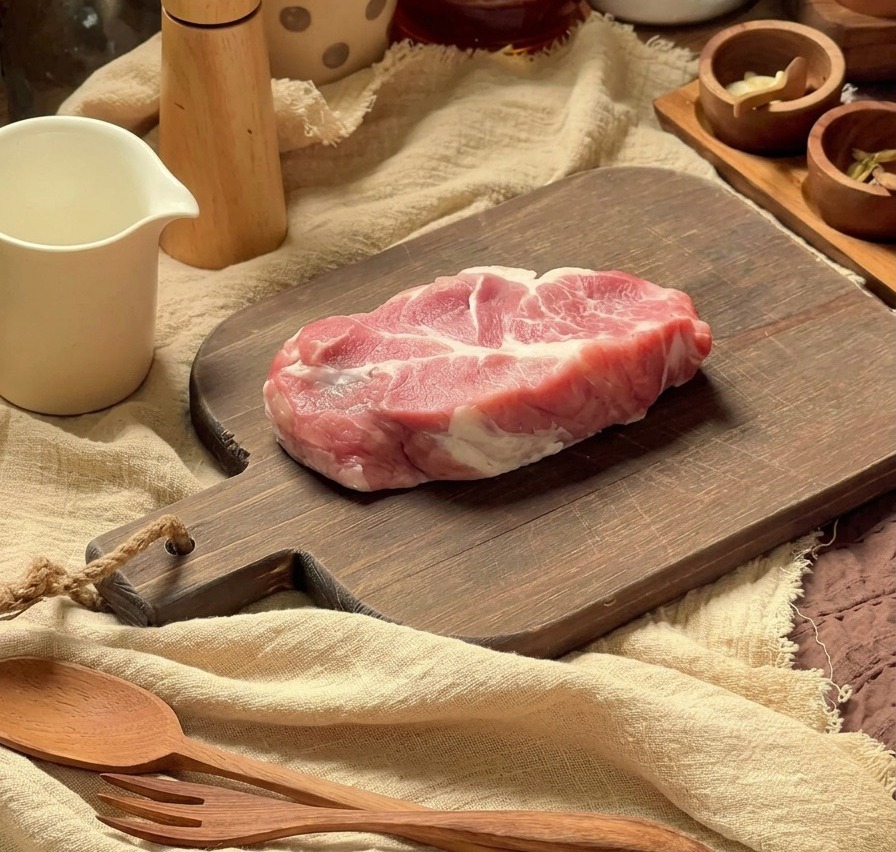 Fetta di coppa di maiale cruda preparata su un tagliere di legno con utensili da cucina sullo sfondo.