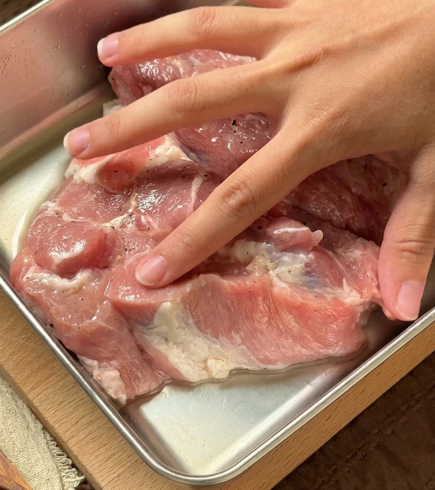 Una mano che massaggia un liquido aromatico trasparente sulle fette di maiale allinterno di un contenitore metallico.