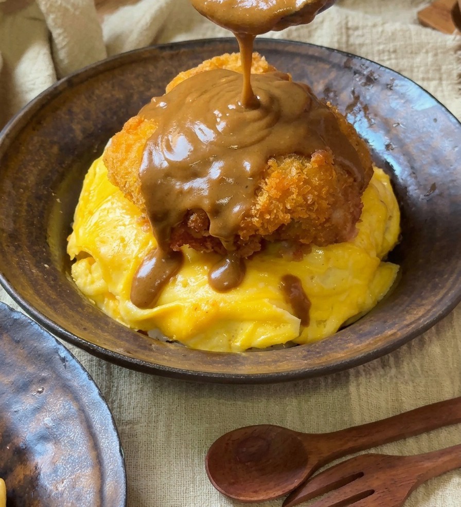 Una salsa al curry marrone e densa versata da un cucchiaio di legno su una cotoletta dorata adagiata su uova a spirale.
