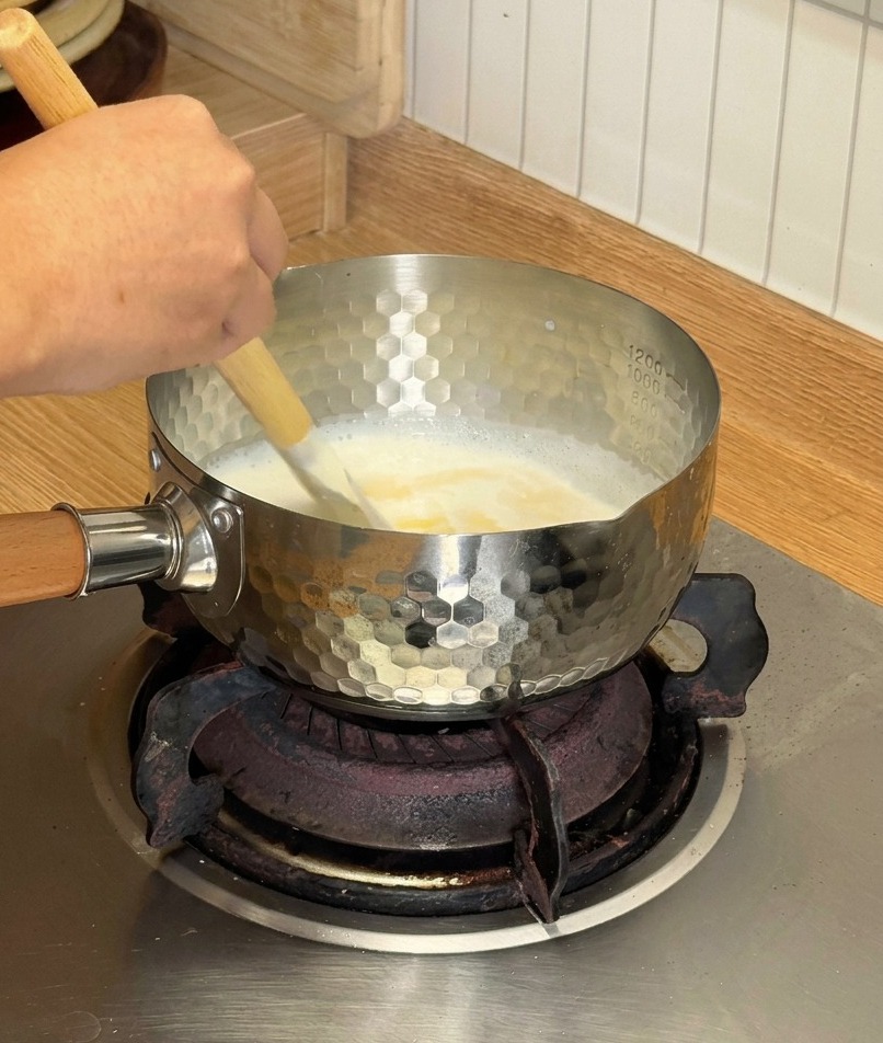 Una spatola di legno che mescola un composto cremoso di formaggio fuso e latte di cocco in un pentolino dargento su un fornello a gas.