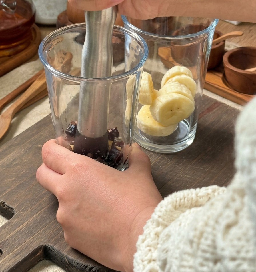Mani che usano un pestello di metallo per schiacciare gli ingredienti in un bicchiere alto, con un secondo bicchiere di banane affettate su un tagliere di legno.