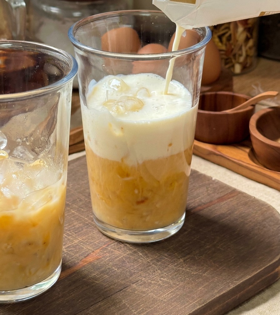 Latte freddo versato da un cartone in un bicchiere alto pieno di ghiaccio e una base di purea di banana gialla.