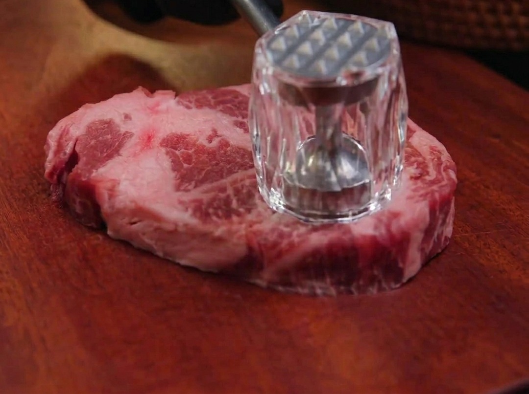 Una mano usa un batticarne di metallo per appiattire una fetta di spalla di maiale cruda su un tagliere di legno scuro.