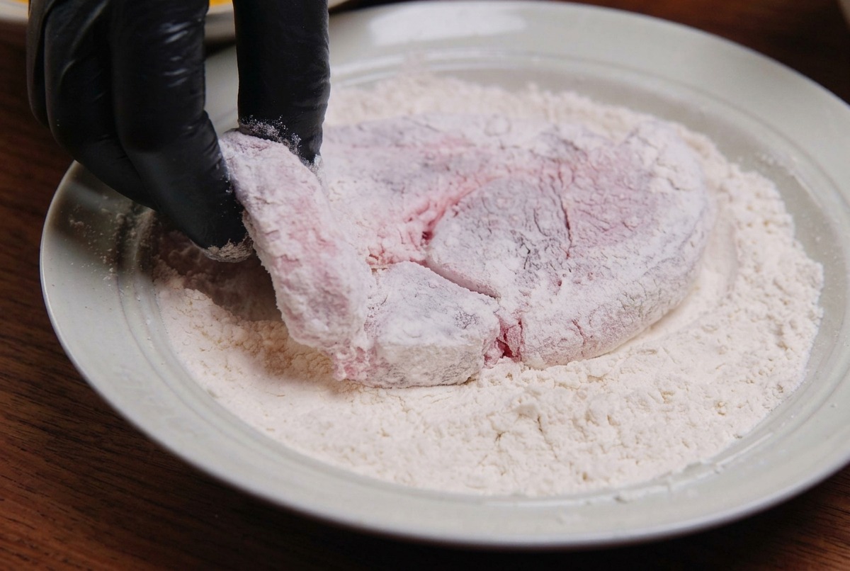 Una fetta di maiale crudo viene passata nella farina bianca allinterno di un piatto di ceramica.