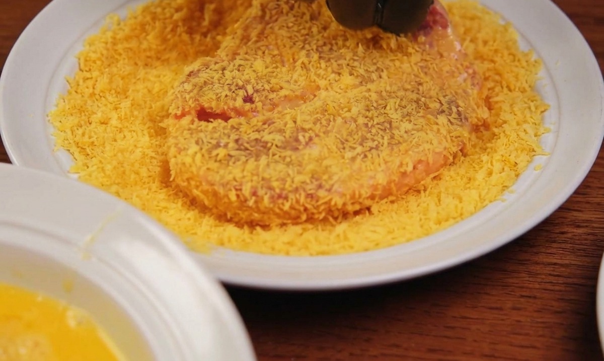 Una mano che indossa un guanto nero preme un pezzo di carne su un piatto ricoperto di panko dorato.
