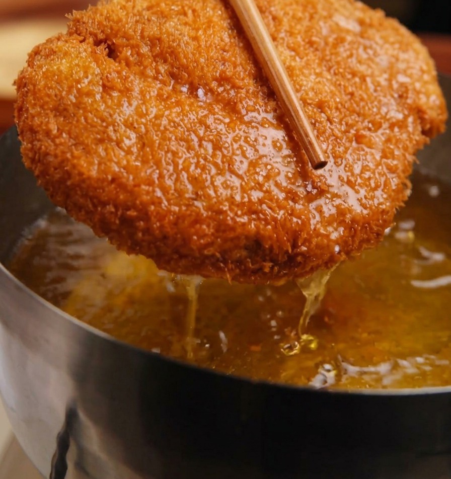 Una cotoletta di maiale dorata e impanata trattenuta con delle bacchette di legno sopra una pentola di olio sfrigolante.