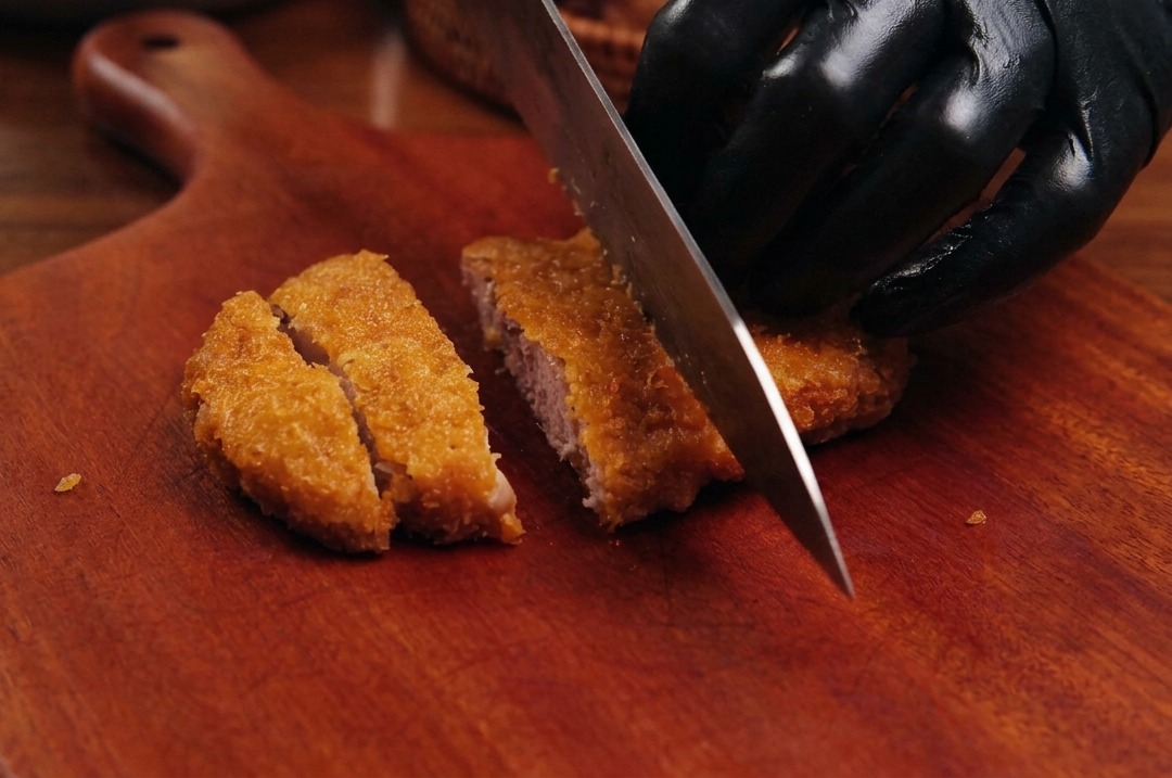 Un coltello da chef affilato taglia una cotoletta fritta dorata e impanata su un tagliere di legno.
