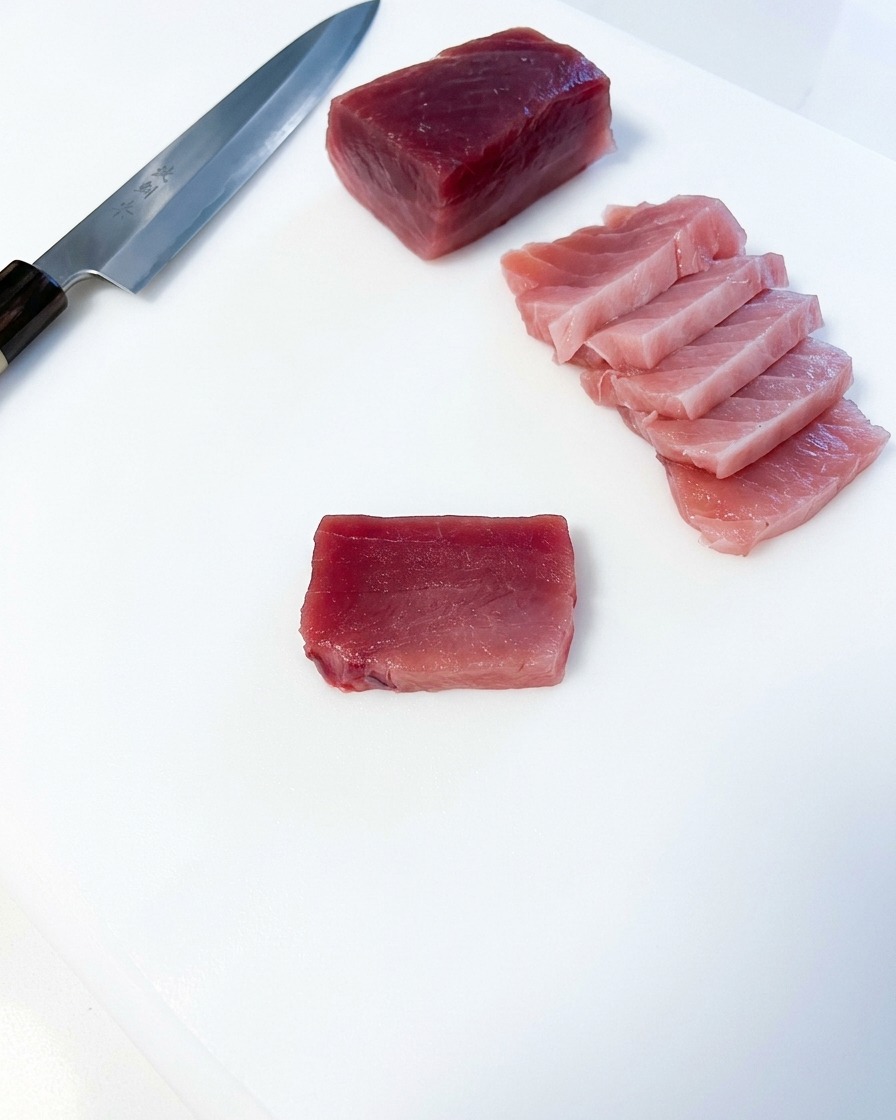 Diverse fette rettangolari di tonno grasso rosa disposte accanto a un grande blocco di tonno e a un coltello da cucina giapponese su un tagliere bianco.