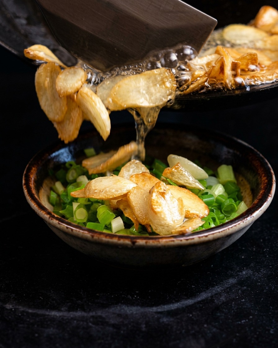 Olio bollente sfrigolante e scaglie di aglio fritto versati da una padella in una ciotola di cipollotti tritati.