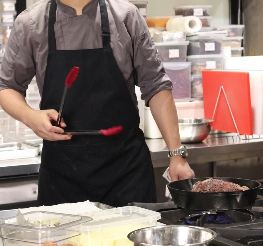 Uno chef che usa le pinze sopra una padella calda in ghisa contenente un grande pezzo di manzo per scottare il grasso.