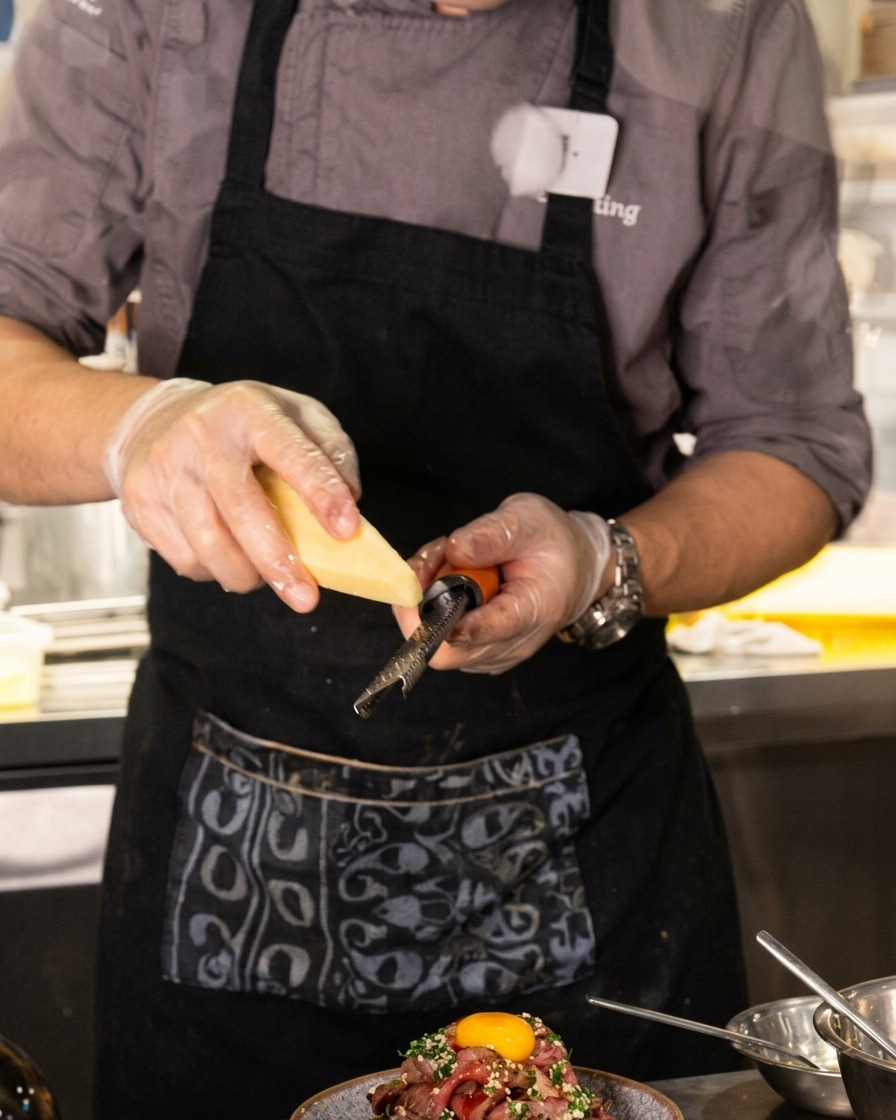 Un primo piano dello chef che usa una grattugia microplane per ridurre a scaglie sottili il formaggio sopra una montagna di manzo con un tuorlo duovo in cima.