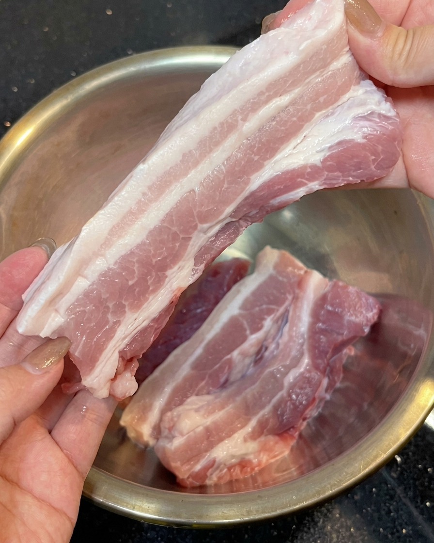 Due mani che tengono una lunga striscia di pancetta di maiale cruda a strati rosa e bianchi con la cotenna rimossa, sopra una ciotola di acciaio inossidabile.