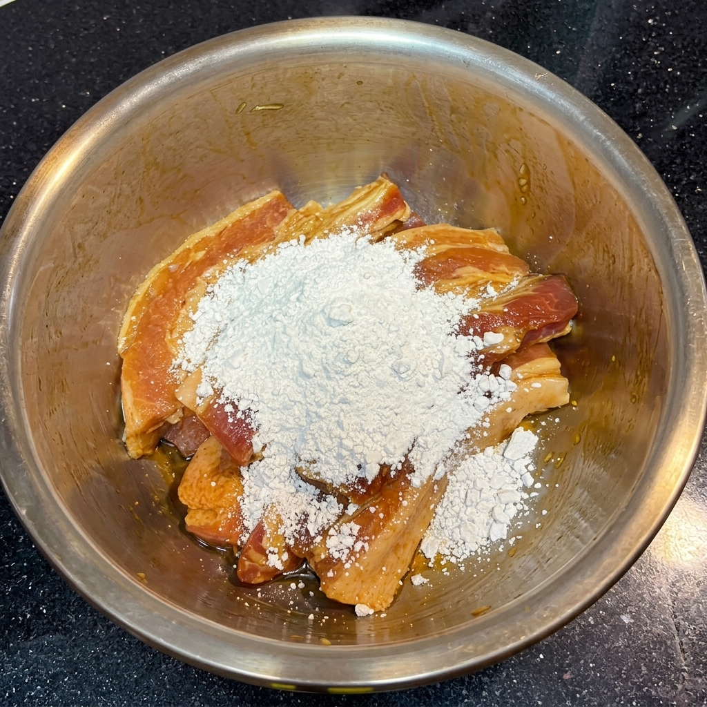 Una generosa manciata di amido di patate dolci bianco e farinoso sopra strisce di pancetta di maiale marinate marrone scuro in una ciotola.
