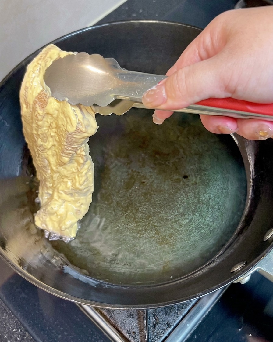 Un pezzo di pancetta di maiale pastellata delicatamente immerso nellolio caldo in padella usando delle pinze di metallo.
