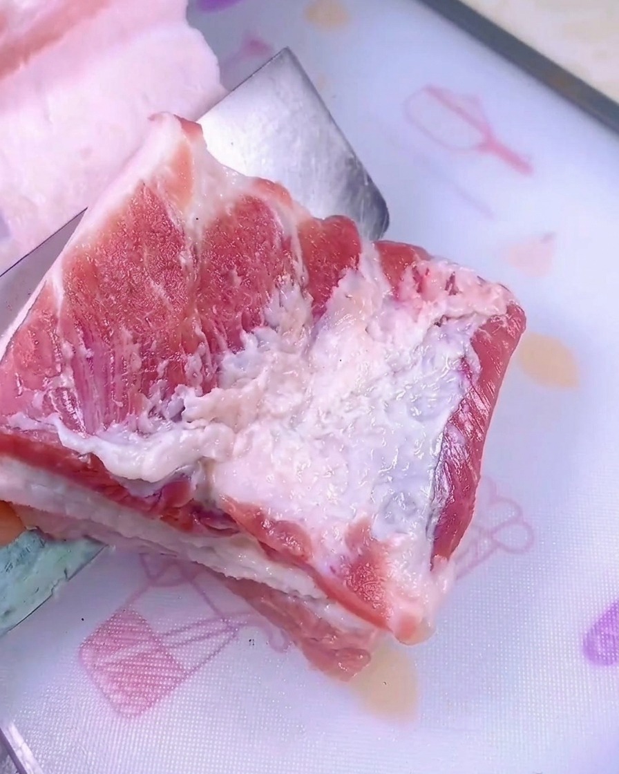 Un coltello affilato taglia un pezzo di pancetta di maiale cruda su un tagliere di plastica bianco.