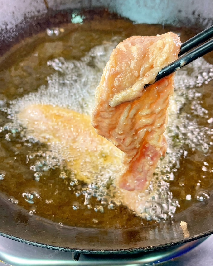 Una fetta di pancetta di maiale pastellata viene immersa nellolio bollente con delle bacchette nere.