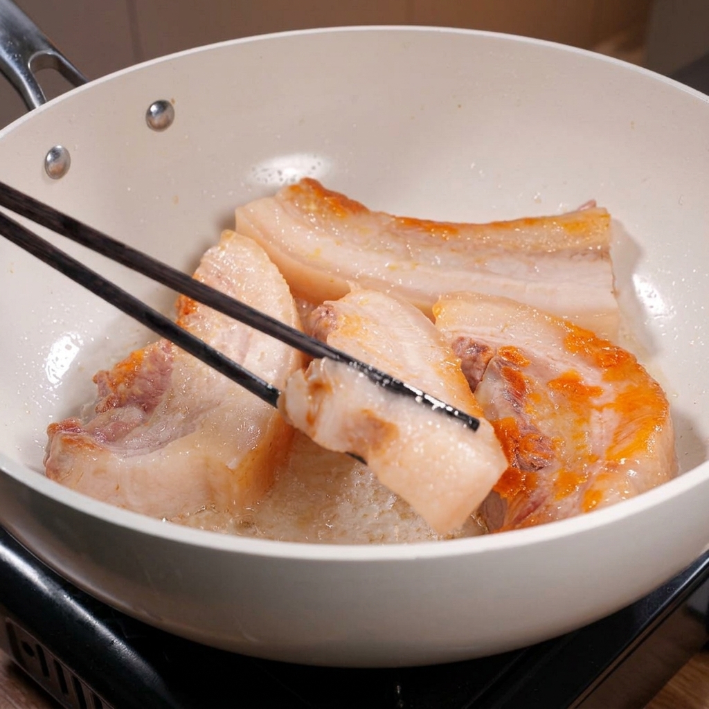 Lunghe bacchette nere girano spesse strisce di pancetta di maiale in una padella bianca, mostrando bordi splendidamente rosolati e dorati.