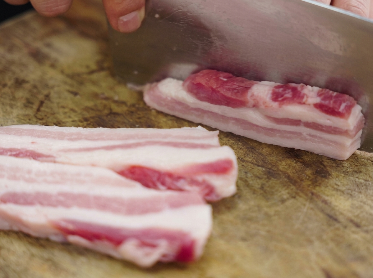 Spesse strisce di pancetta di maiale cruda a strati tagliate su un tagliere di legno con una grande mannaia di metallo.