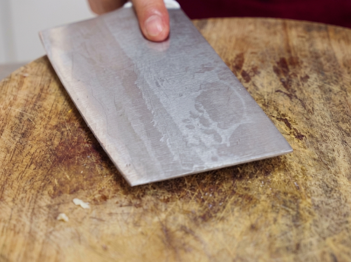 Una pesante mannaia di metallo premuta piatta su un tagliere di legno per schiacciare spicchi daglio crudo.
