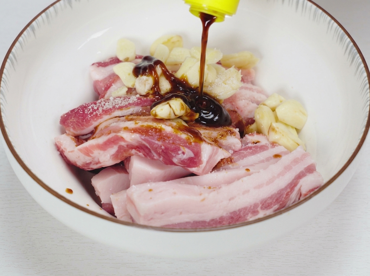 Condimenti liquidi scuri e salsa dostriche versati sulla pancetta di maiale cruda a fette e aglio schiacciato in una ciotola di ceramica bianca.
