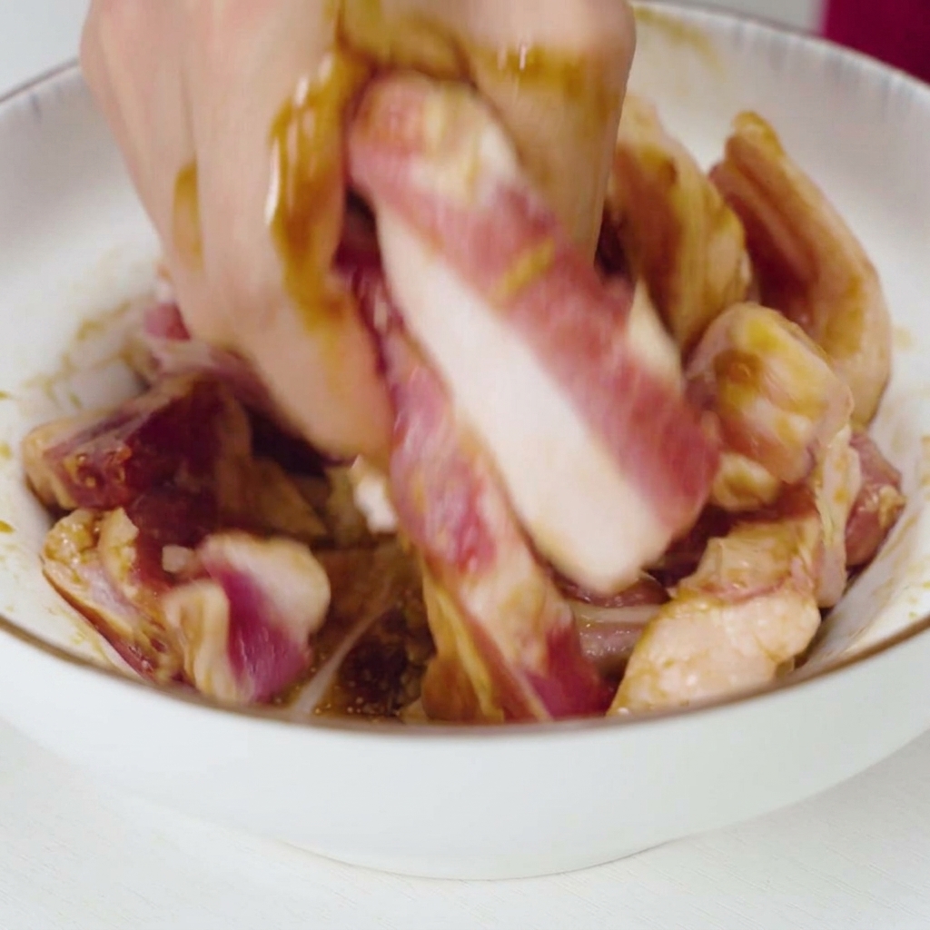 Una mano che mescola e massaggia energicamente strisce di maiale crudo con una marinatura liquida scura e pezzi daglio in una ciotola di ceramica bianca.