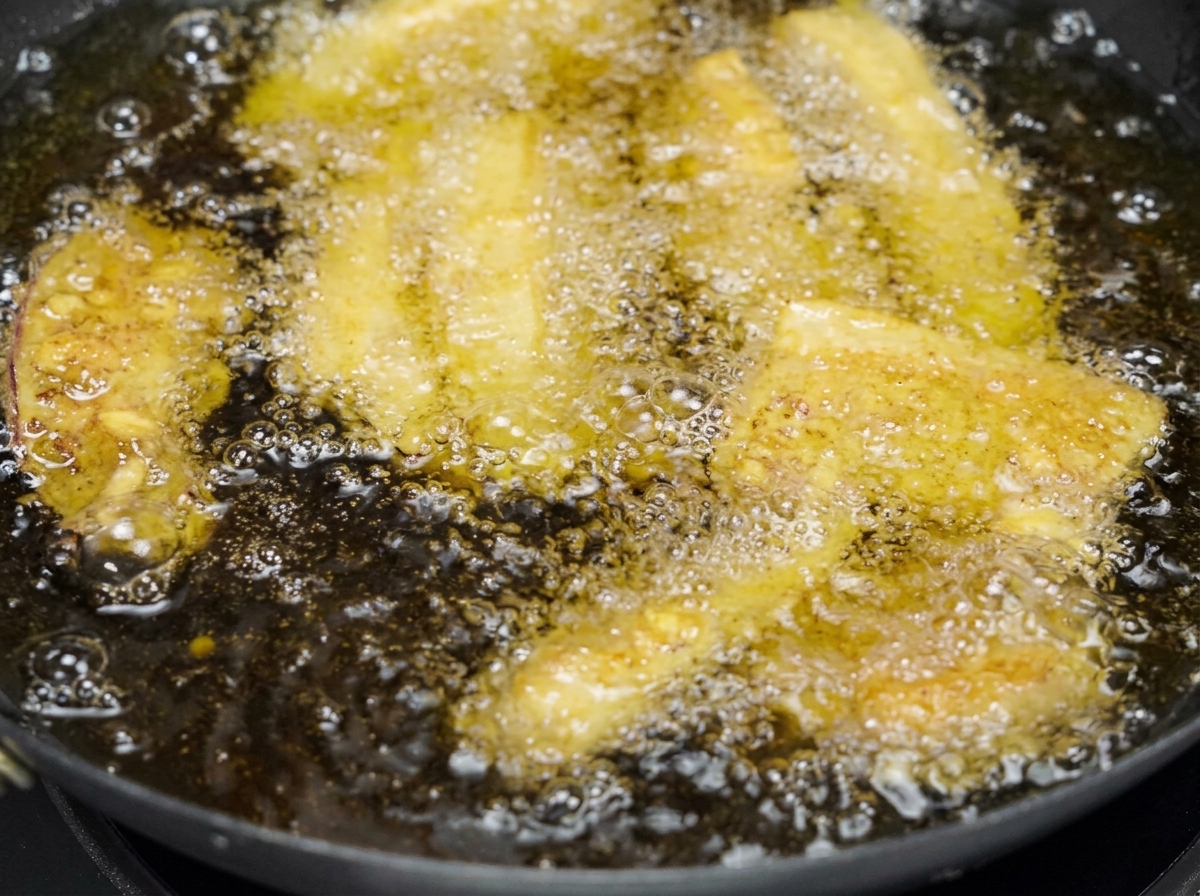 Diverse strisce di pancetta di maiale impanata che friggono lentamente in una padella piena di olio dorato e spumeggiante.