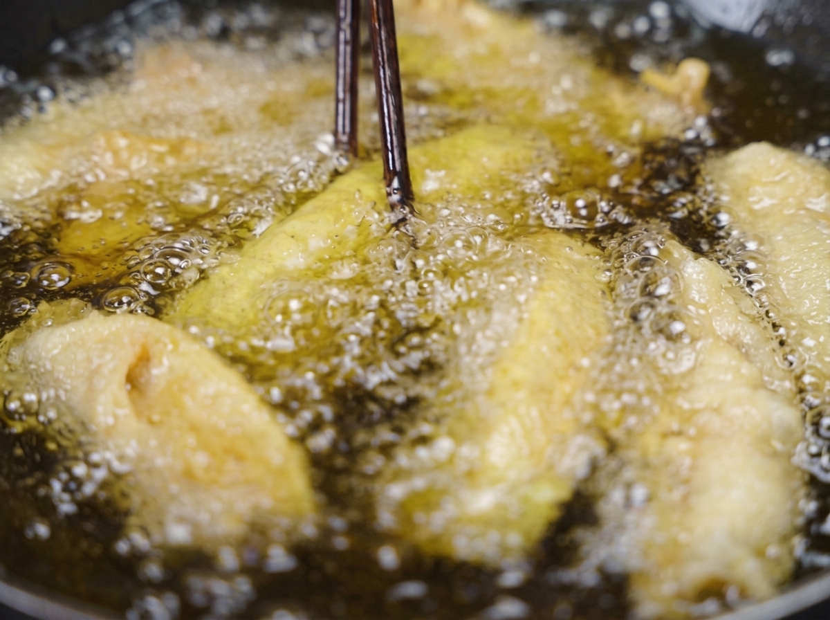 Bacchette di legno scuro che sollevano una striscia di maiale fritto croccante e dorato dallolio bollente.