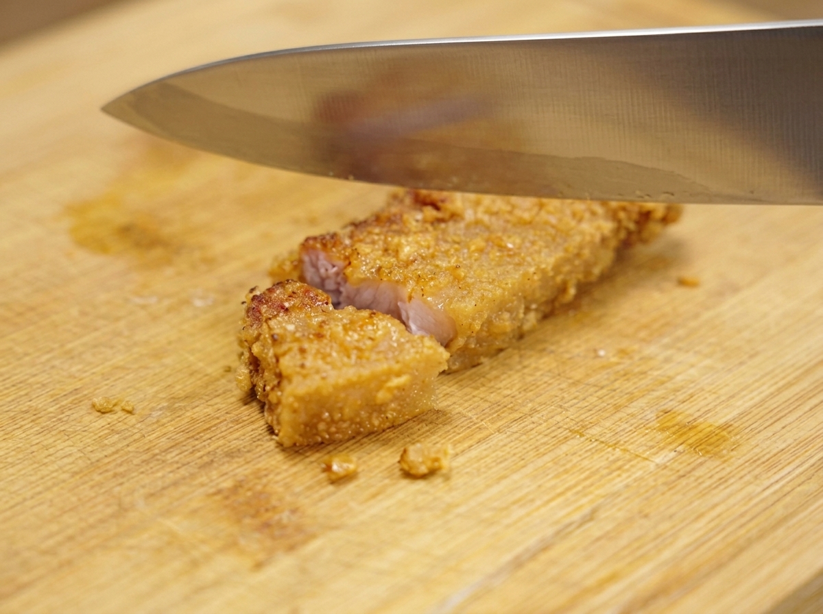 Un grande coltello di metallo che taglia un pezzo croccante e dorato di maiale fritto in porzioni più piccole su un tagliere di legno.
