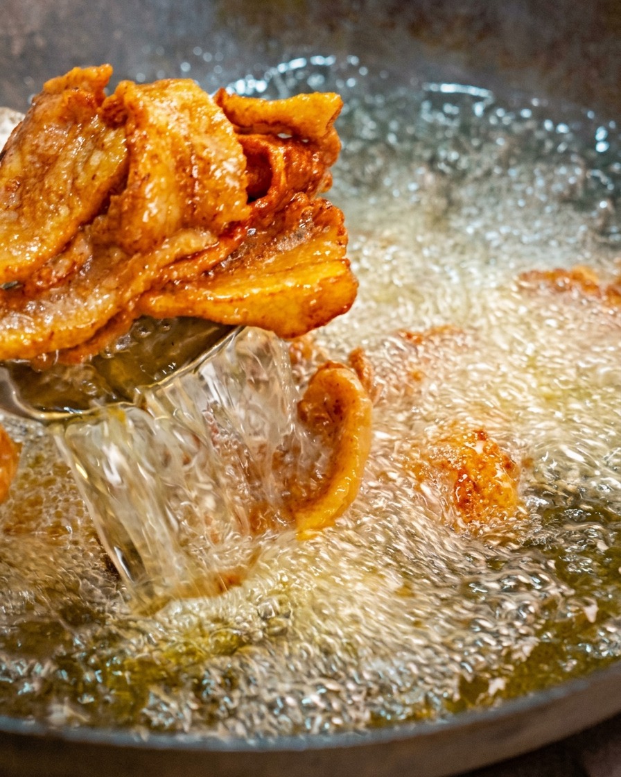 Prelevare fette dorate e arricciate di pancetta di maiale fritta da un wok di olio bollente usando una schiumarola a rete.