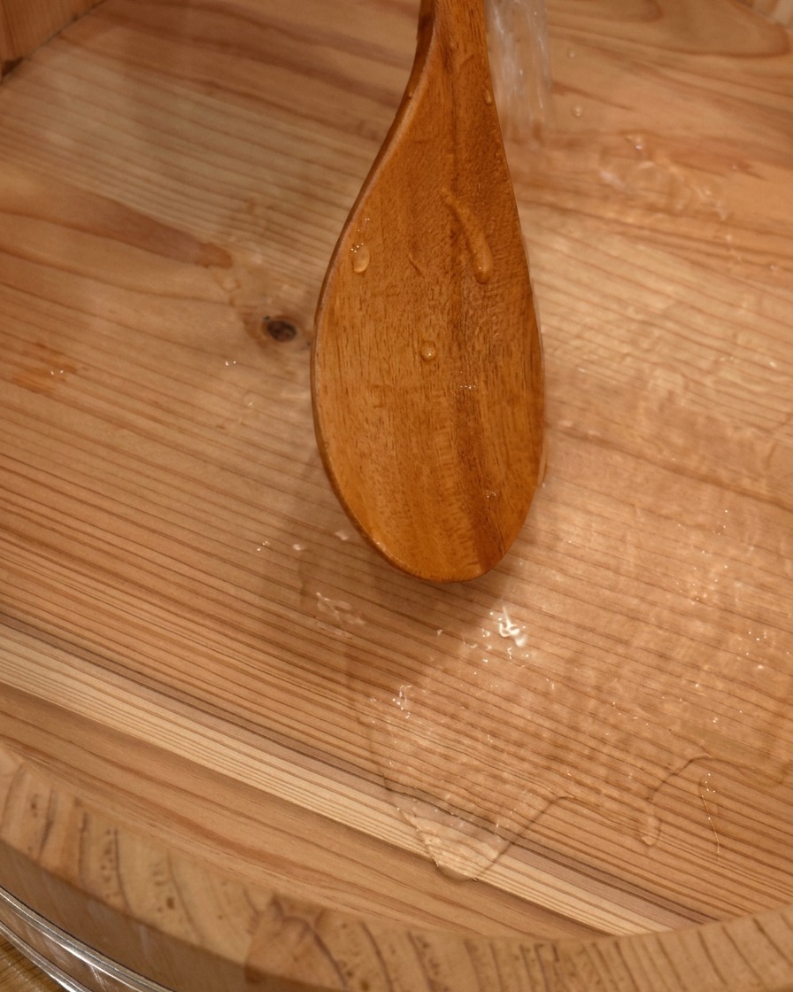 Acqua che viene versata da una paletta di legno in un grande contenitore di legno tradizionale per mescolare il riso del sushi.
