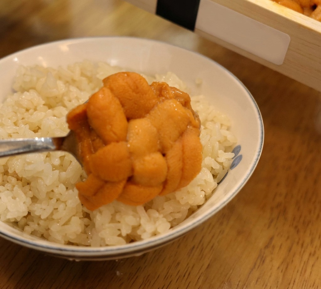 Un cucchiaio che solleva una polpa di riccio di mare arancione per adagiarla su una ciotola di riso bianco per sushi.